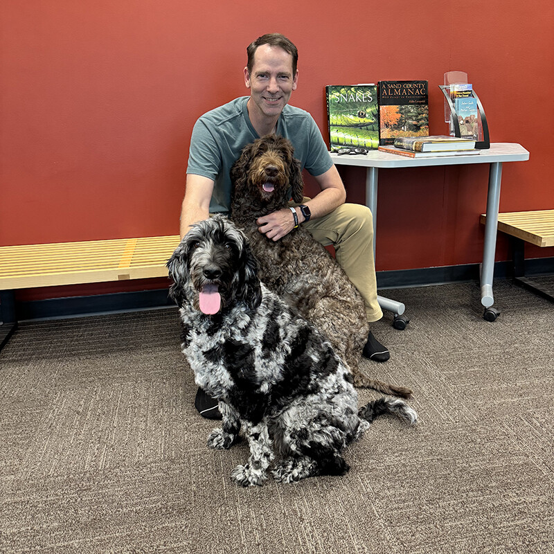 Kenneth Pyle sits in a blue shirt and khaki pants with an arm around Jonas, a brown and white labradoodle, with River, a black and grey labradoodle, also sitting in front of them.
