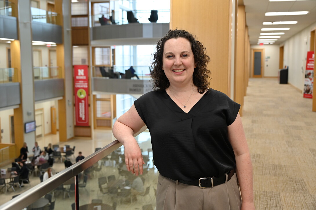 Rebecca Jack, a white woman with long, curly dark hair stands in a black blouse and tan trousers in the College of Business.