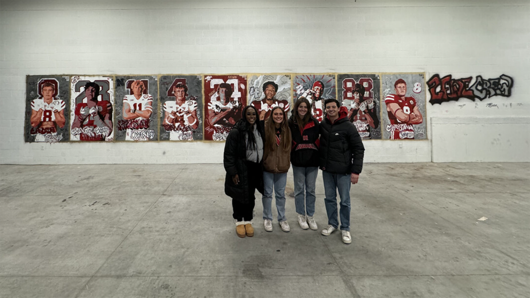 Art students stand in an big open room with the Husker recruit murals painted on the wall behind them.