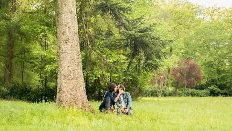 A man with brown hair and a beard, dressed in a white shirt and jean jacket, kisses a woman with brown hair, in a dark blue shirt, next to a tree in a clearing in a forest.