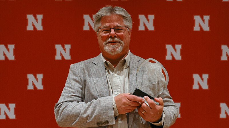Mark Vrtiska stands in a grey suit in front of a red backdrop, with short grey hair and thinly framed glasses. He is smiling and holding an award.