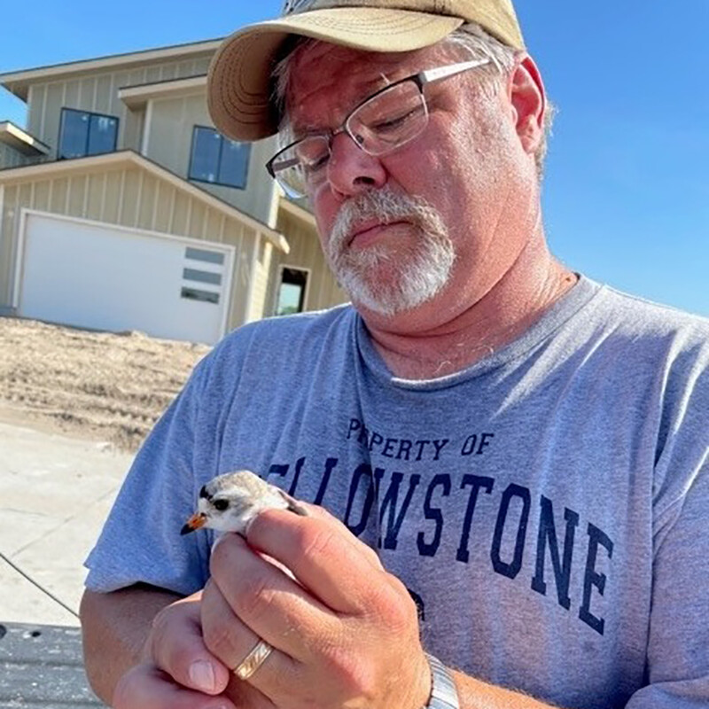 Mark Vrtiska stands in front of a tan building wearing a grey t-shirt and a tan hat, with thin-frame glasses on, holding a small white and grey bird with an orange beak.