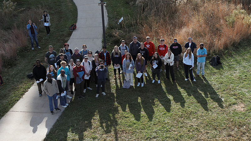 A class of people stand together outside, smiling while looking up.