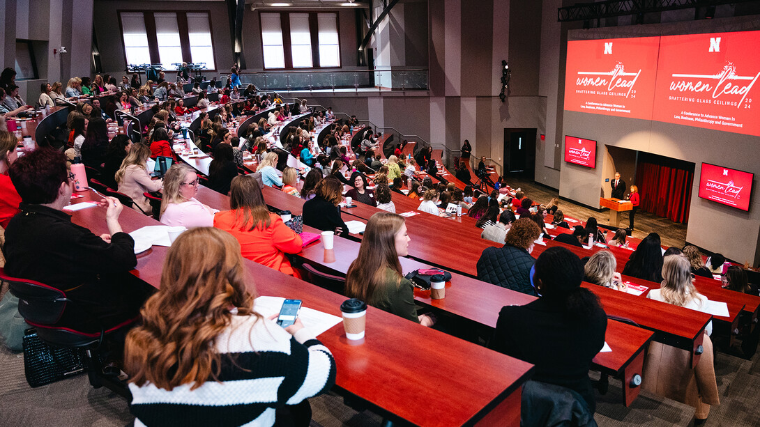 A crowd of people at red tables watch a woman present in front of a projector at a past Women Lead conference.