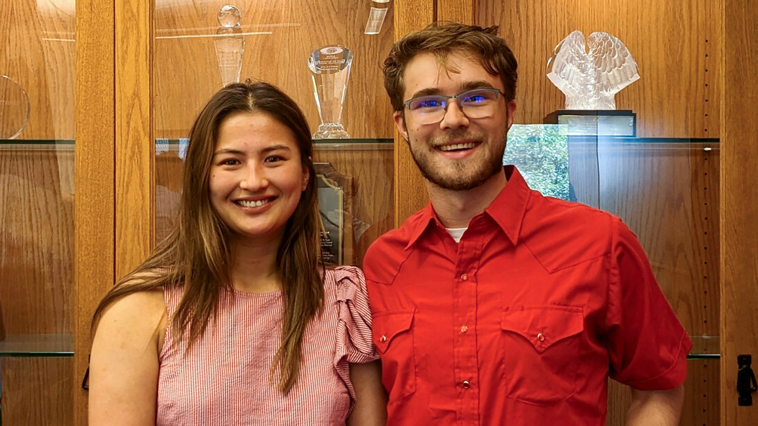 Kyoko Wall stands on the left in a pink blouse, smiling with long brown hair, and Zane Mrozla-Mindrup stands on the right, smiling in a red button-up shirt with glasses, short brown hair and a beard. Both stand in front of a wooden cabinet with glass awards in it.