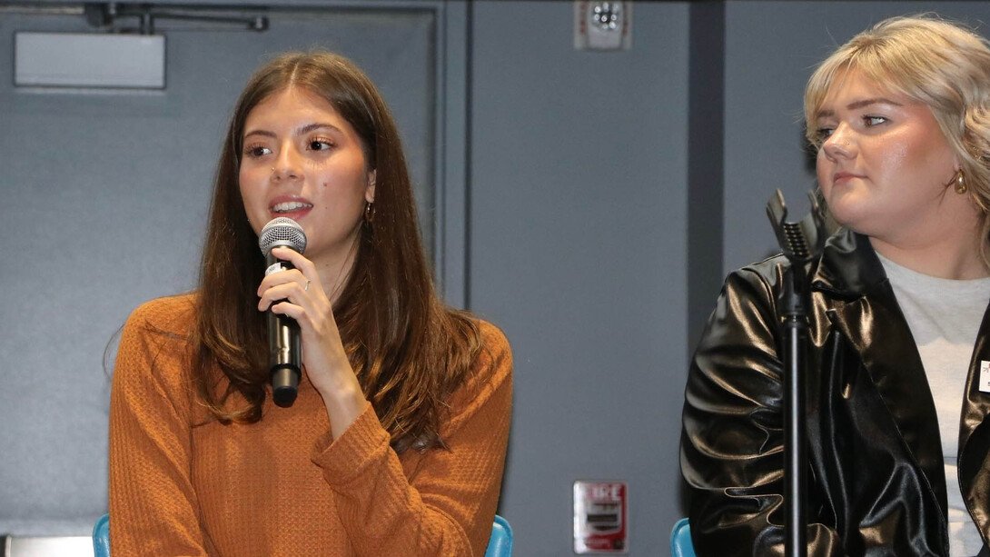 A brunette woman in an orange shirt speaks into a microphone while sitting next to a blonde woman in a white sweater and black leather jacket.