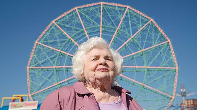 An old woman stands in a pink shirt and jacket with a large ferris wheel in the background.