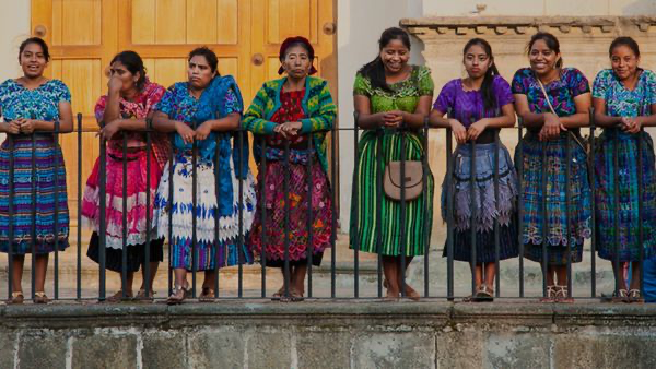 Women in colorful dresses standing along a fence.