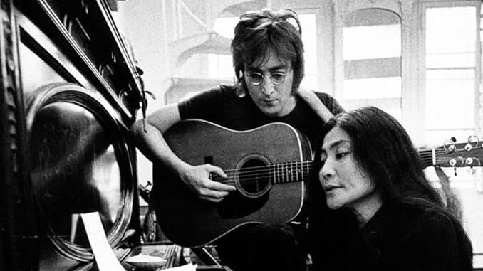 John Lennon plays guitar next to Yoko Ono, who is playing a piano, in a black and white photo.