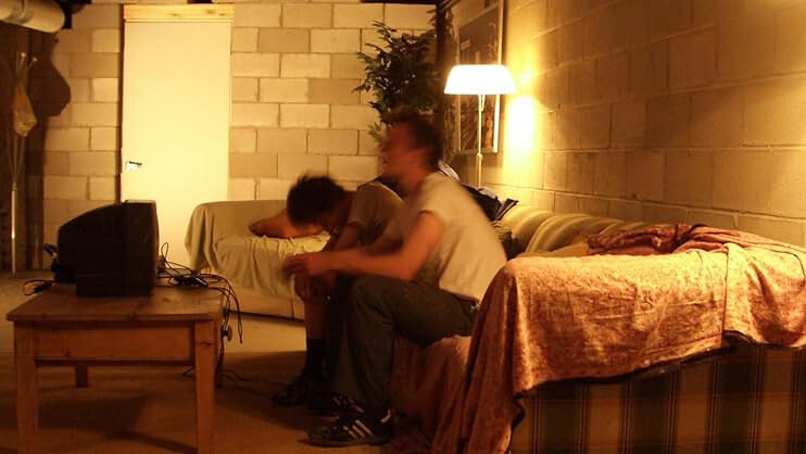 Two people sit on a couch, in front of a coffee table with a small tv, in a room with concrete brick walls.