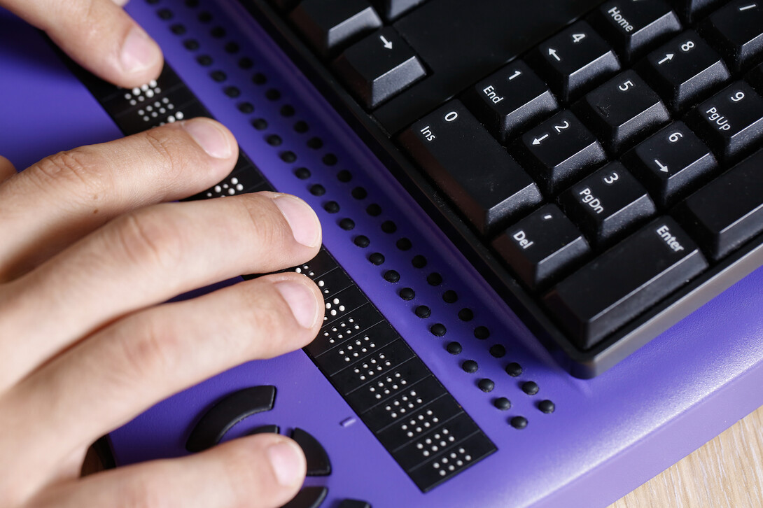 A person uses a computer with braille display and a computer keyboard.
