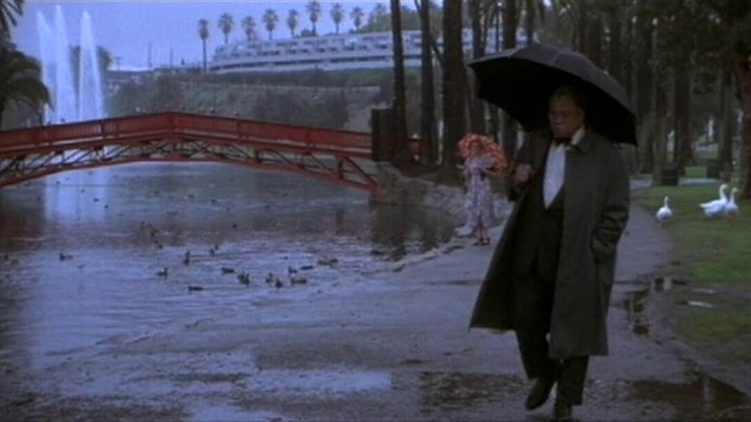 A man in a black suit and coat holds an umbrella while walking along a riverbank.